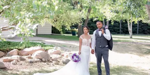 A bride and groom standing outdoors in a garden setting, holding hands and posing for a wedding photograph.