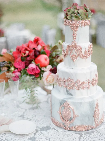A four-tier wedding cake with a white marble fondant finish, decorated with intricate rose-gold sugar ornaments and topped with fresh flowers, displayed on a table with a floral arrangement.
