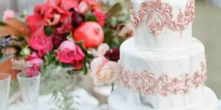 A four-tier wedding cake with a white marble fondant finish, decorated with intricate rose-gold sugar ornaments and topped with fresh flowers, displayed on a table with a floral arrangement.