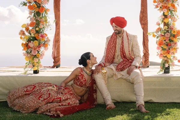 A bride and groom in traditional Indian wedding attire posing together at a ceremony decorated with vibrant floral arrangements.