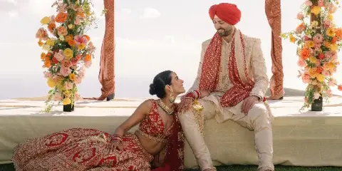 A bride and groom in traditional Indian wedding attire posing together at a ceremony decorated with vibrant floral arrangements.