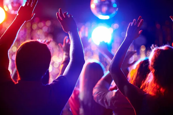 A crowd of people dancing with hands raised under colorful stage lights and a disco ball.