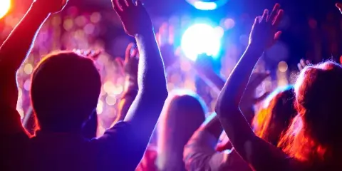 A crowd of people dancing with hands raised under colorful stage lights and a disco ball.