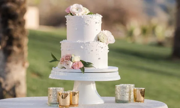 A white two-tier wedding cake decorated with fresh flowers and pearl accents, displayed on a white pedestal stand outdoors.