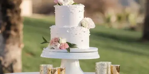 A white two-tier wedding cake decorated with fresh flowers and pearl accents, displayed on a white pedestal stand outdoors.
