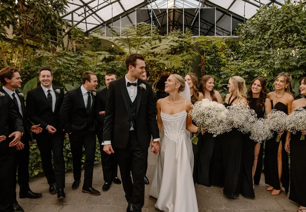 A bride and groom walking hand-in-hand through a lush greenhouse, surrounded by their wedding party.