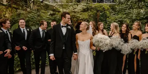 A bride and groom walking hand-in-hand through a lush greenhouse, surrounded by their wedding party.