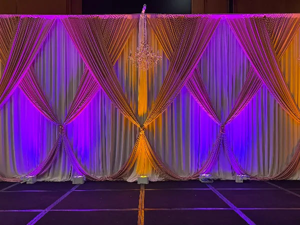 An event stage backdrop featuring draped white linens, a crystal chandelier, and purple and orange uplighting.