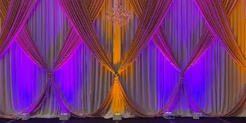 An event stage backdrop featuring draped white linens, a crystal chandelier, and purple and orange uplighting.