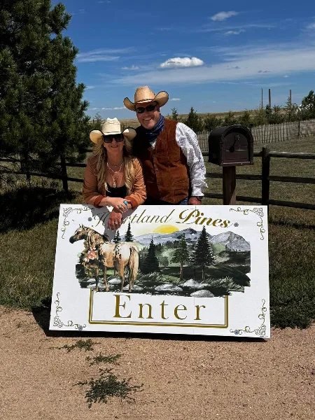 Two people in cowboy hats standing behind a painted wooden sign that reads 'Heartland Pines Enter' with a horse and mountain illustration.