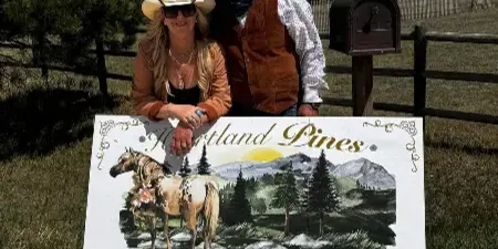 Two people in cowboy hats standing behind a painted wooden sign that reads 'Heartland Pines Enter' with a horse and mountain illustration.