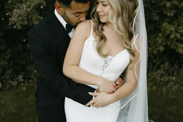 A groom in a black tuxedo embraces a bride in a white lace dress and veil against a dark, leafy background.