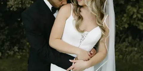 A groom in a black tuxedo embraces a bride in a white lace dress and veil against a dark, leafy background.