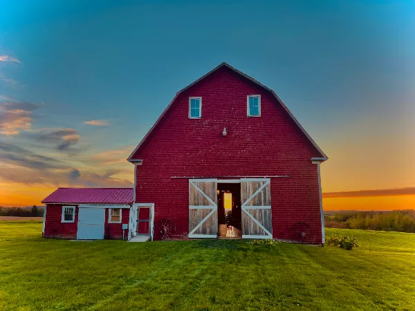 A red wooden barn with open sliding doors set against a sunset sky in a green field.