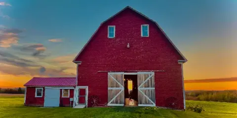 A red wooden barn with open sliding doors set against a sunset sky in a green field.