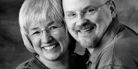 A black and white portrait of a smiling woman and man, identified as the photographers Sharon and Greg Lewis.