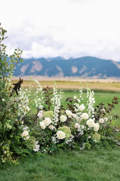A large outdoor floral arrangement featuring white roses and greenery set against a backdrop of a grassy field and mountains.