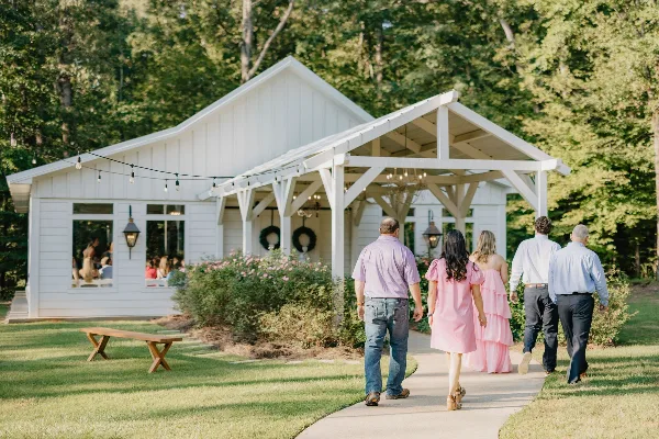 Guests walking on a paved path toward a white chapel and reception building with a covered porch, surrounded by green lawn and trees.