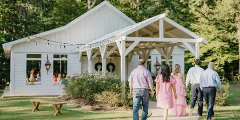 Guests walking on a paved path toward a white chapel and reception building with a covered porch, surrounded by green lawn and trees.