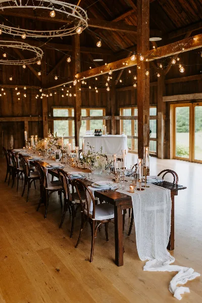 Interior of a rustic barn venue with a long wooden table set for a wedding reception, decorated with candles, floral centerpieces, and hanging string lights.