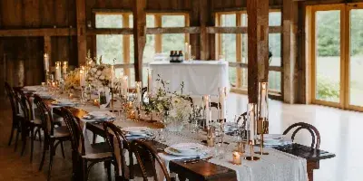 Interior of a rustic barn venue with a long wooden table set for a wedding reception, decorated with candles, floral centerpieces, and hanging string lights.