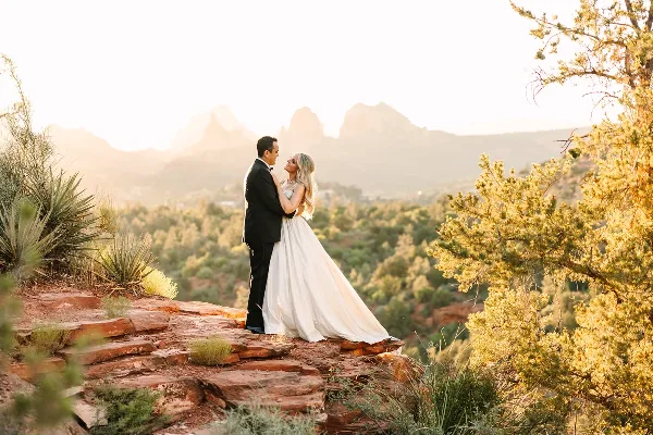 A bride and groom standing on a red rock ledge with a view of Sedona's desert landscape and mountains in the background.