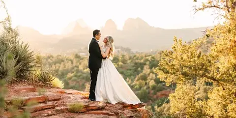 A bride and groom standing on a red rock ledge with a view of Sedona's desert landscape and mountains in the background.
