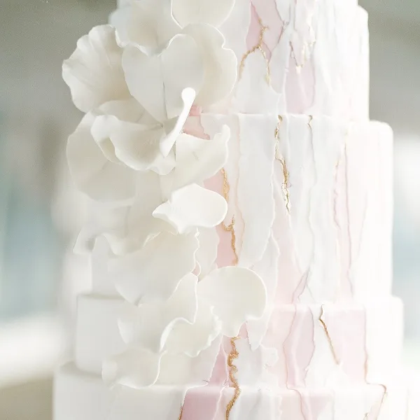 A close-up of a white and pink multi-tiered wedding cake decorated with white fondant flowers and gold accents.