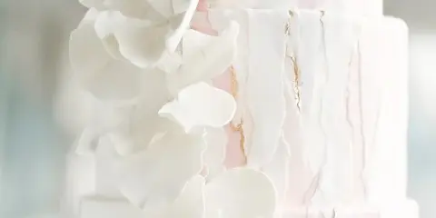 A close-up of a white and pink multi-tiered wedding cake decorated with white fondant flowers and gold accents.