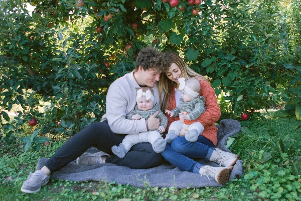 A family of four sitting on a blanket in an apple orchard, with a man and woman holding two babies wearing matching headbands.