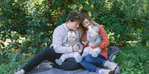 A family of four sitting on a blanket in an apple orchard, with a man and woman holding two babies wearing matching headbands.