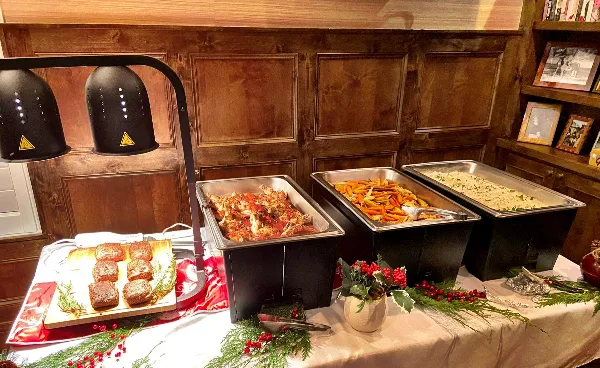A catering buffet setup featuring chafing dishes with prepared food, heat lamps, and a plated side dish on a decorated table.