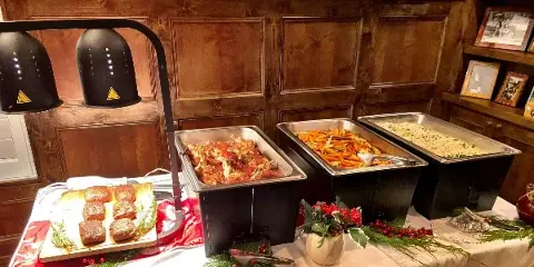 A catering buffet setup featuring chafing dishes with prepared food, heat lamps, and a plated side dish on a decorated table.