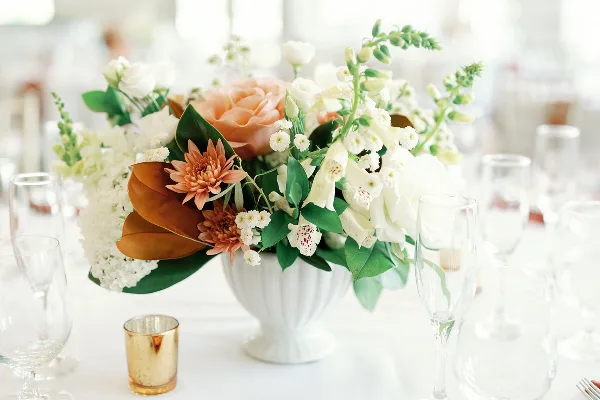 A floral centerpiece featuring white and peach flowers in a white vase on a table setting.