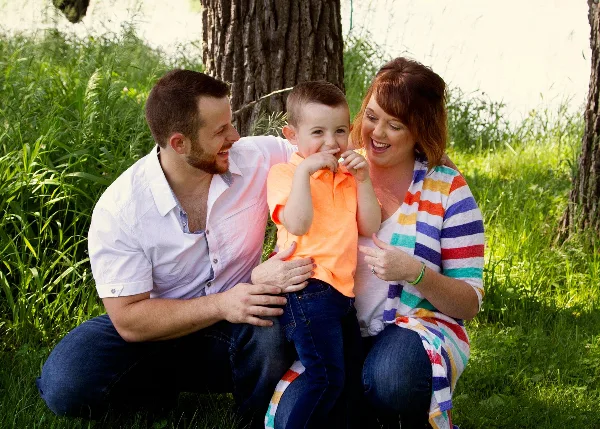 A professional outdoor family portrait of a father, mother, and young child sitting together in a grassy area near a tree.