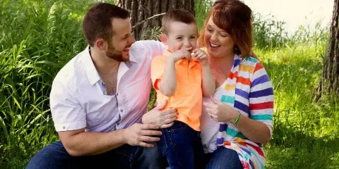 A professional outdoor family portrait of a father, mother, and young child sitting together in a grassy area near a tree.