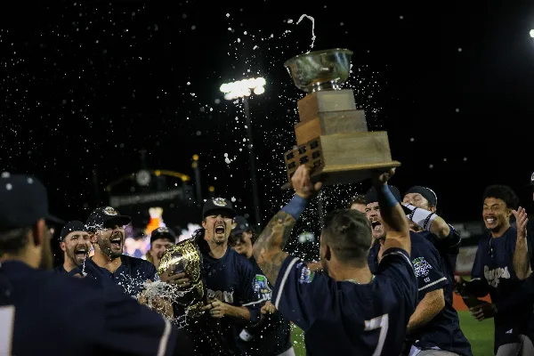 Kane County Cougars baseball players celebrating with a championship trophy on the field at night.