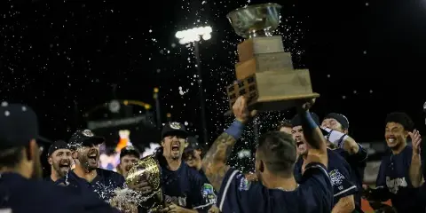 Kane County Cougars baseball players celebrating with a championship trophy on the field at night.