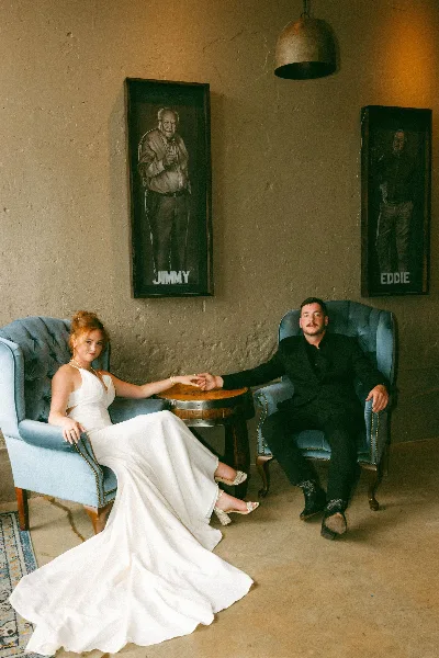 A bride in a white gown and a groom in a black suit sitting in blue armchairs holding hands in a rustic indoor venue.