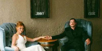 A bride in a white gown and a groom in a black suit sitting in blue armchairs holding hands in a rustic indoor venue.