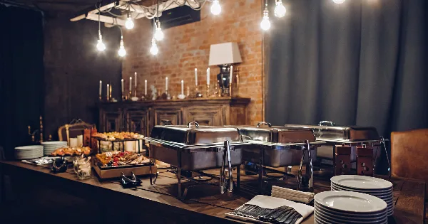 A catering buffet table set with stainless steel chafing dishes, plates, and food trays in an event venue.