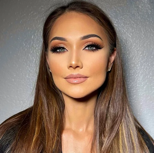 Professional portrait of a woman with long brown hair and full glam makeup, including defined eyebrows, neutral eyeshadow, and glossy lips.
