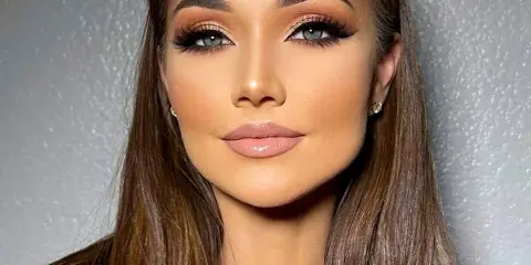Professional portrait of a woman with long brown hair and full glam makeup, including defined eyebrows, neutral eyeshadow, and glossy lips.