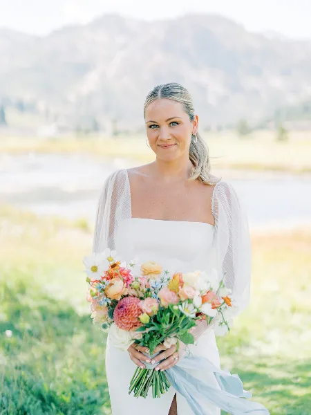 A bride in a white dress holding a colorful bouquet of dahlias and ranunculus against a mountain backdrop.
