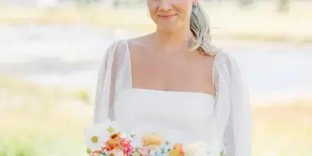 A bride in a white dress holding a colorful bouquet of dahlias and ranunculus against a mountain backdrop.
