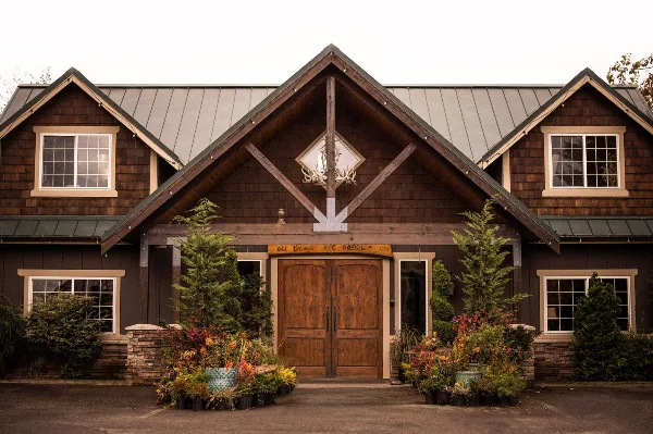 Exterior view of a dark wood barn-style building with a metal roof, featuring a central timber truss, antler decoration, and potted plants in the foreground.