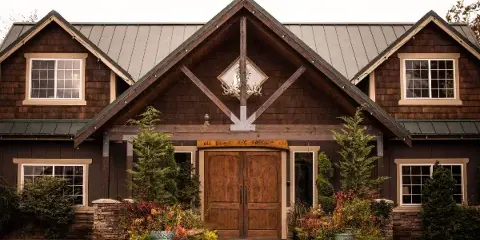 Exterior view of a dark wood barn-style building with a metal roof, featuring a central timber truss, antler decoration, and potted plants in the foreground.