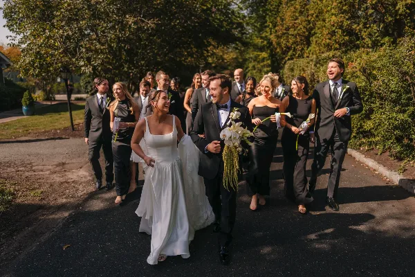 A bride and groom walk down a paved path with their wedding party, captured in a candid documentary style.