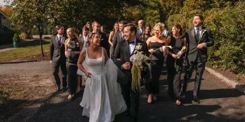 A bride and groom walk down a paved path with their wedding party, captured in a candid documentary style.