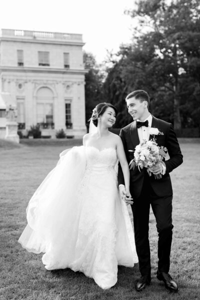 A black and white photograph of a bride and groom walking together on a lawn in front of a large stone building.